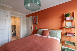 Een slaapkamer met een bed, een bedlamp, een plank met boeken en een deur naar een badkamer.