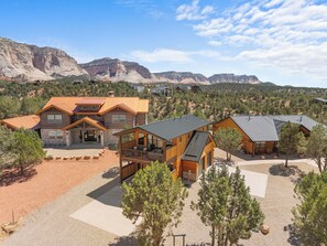 A modern log cabin with a large deck, surrounded by a gravel driveway and a mountainous backdrop. - The Perch – Brand New Zion Retreat with Hot Tub (Orderville)