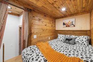 Une pièce aux murs lambrés, avec un lit, un tableau accroché au mur et une fenêtre avec des rideaux.