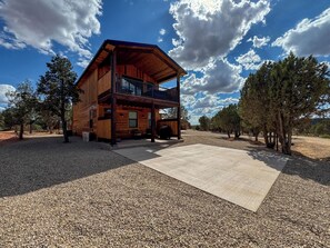 A two-story wooden cabin with a balcony, surrounded by gravel and trees. - The Perch – Brand New Zion Retreat with Hot Tub (Orderville)