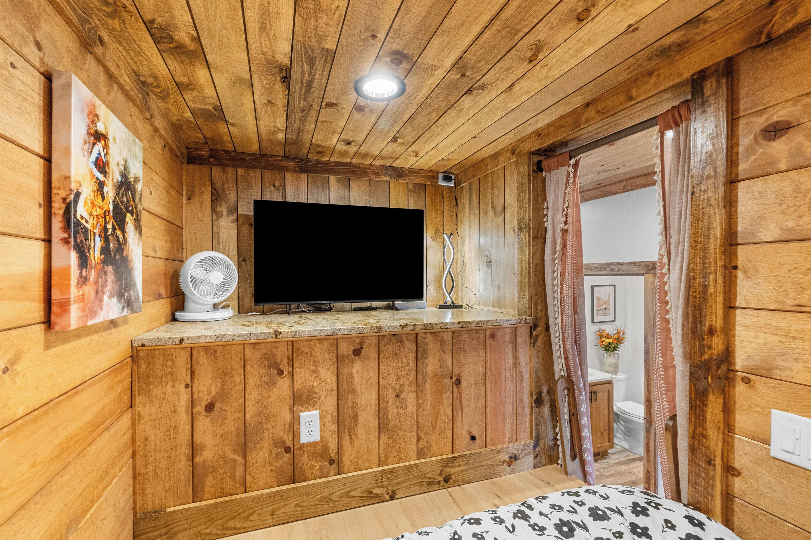 Um quarto com painéis de madeira, uma televisão em uma prateleira, um ventilador e uma cama com estampa floral.