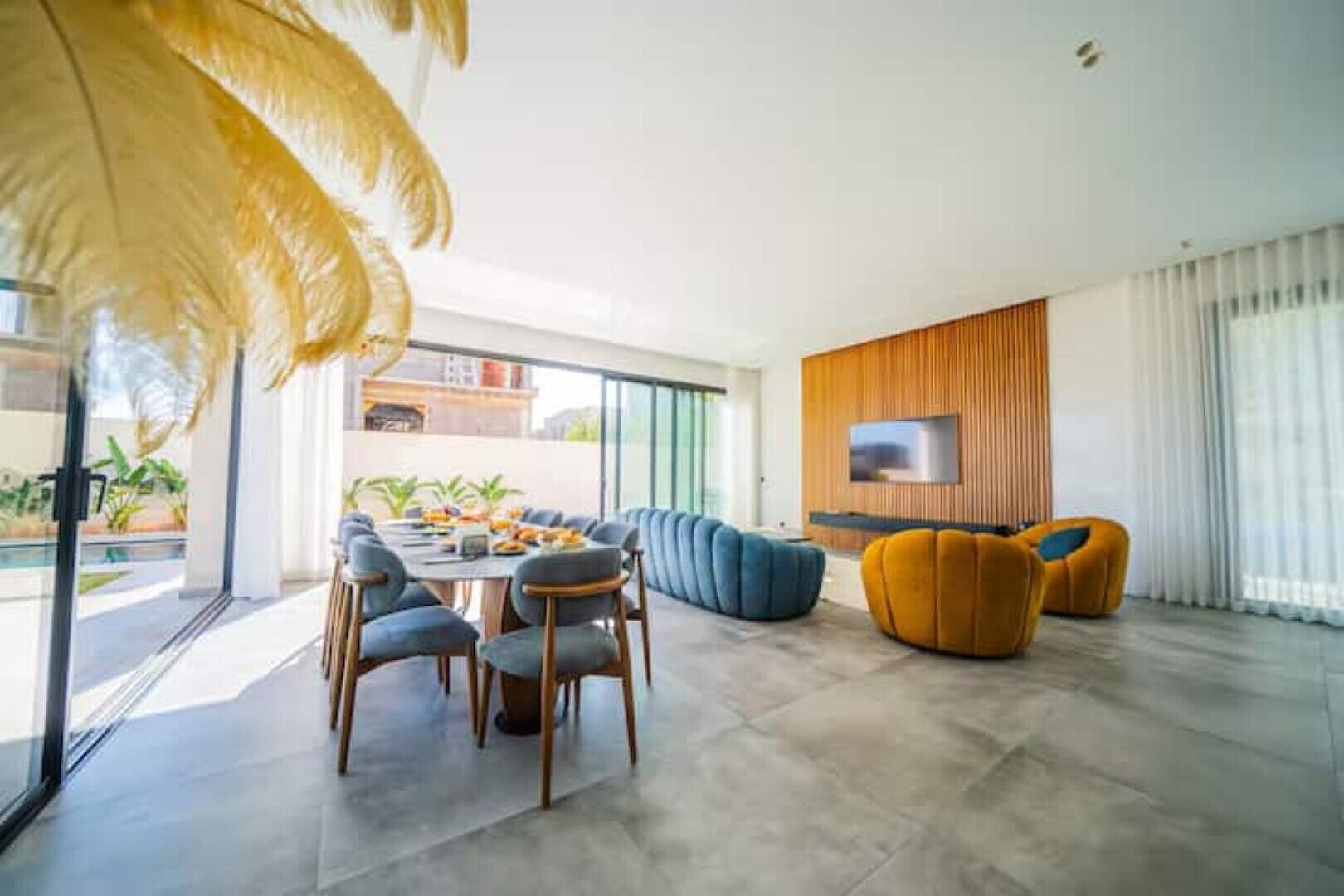 A modern living room with a dining area, a flat-screen TV mounted on a wooden panel, and a large window with a view of the outdoors.