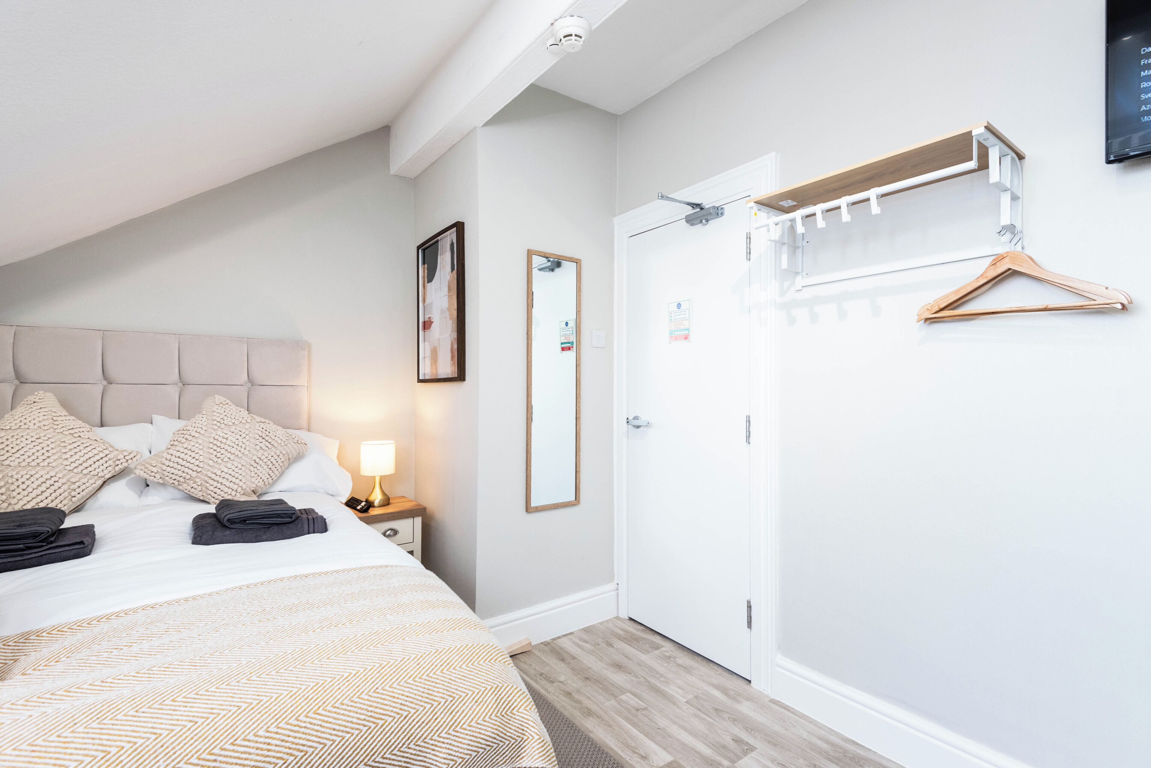 Ein Hotelzimmer mit einem Bett, einem Nachttisch mit Lampe, einem Spiegel, einem Kleiderschrank und einem Garderobenhaken mit Kleiderbügeln.