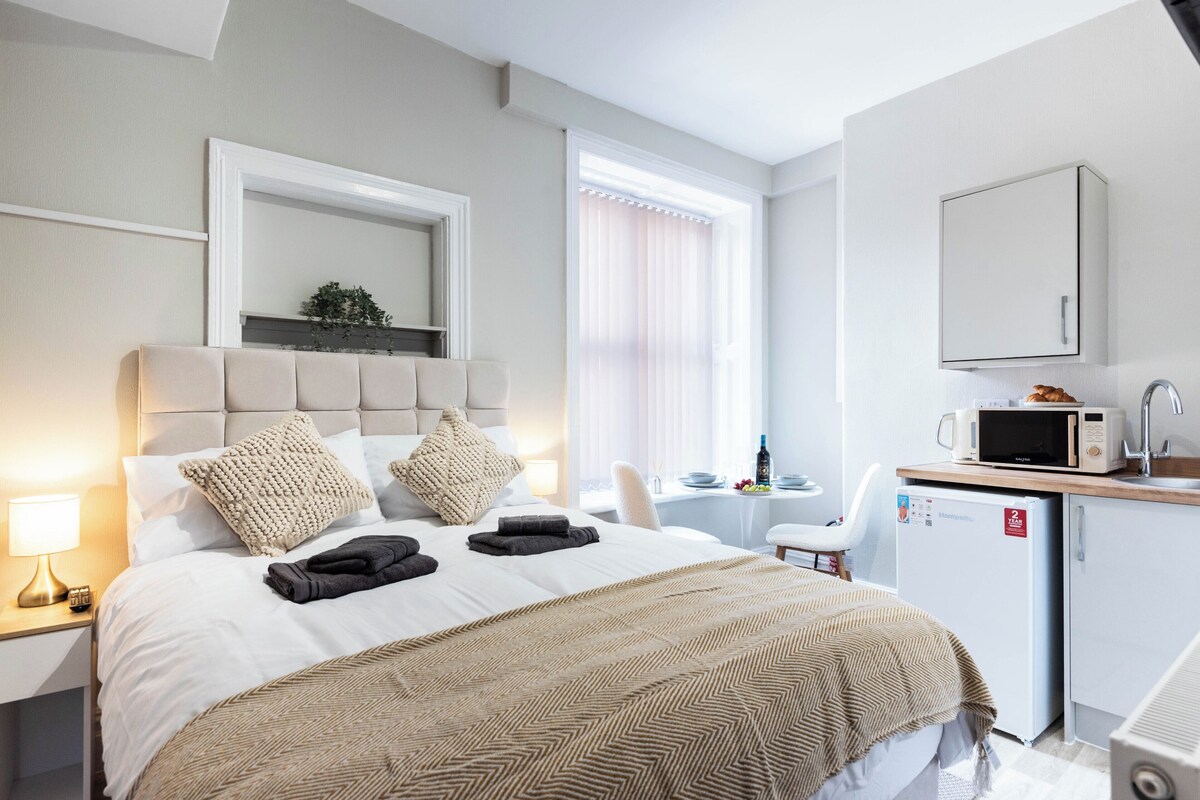 A hotel room with a bed, bedside table, lamp, a small kitchenette with a microwave, and a chair.