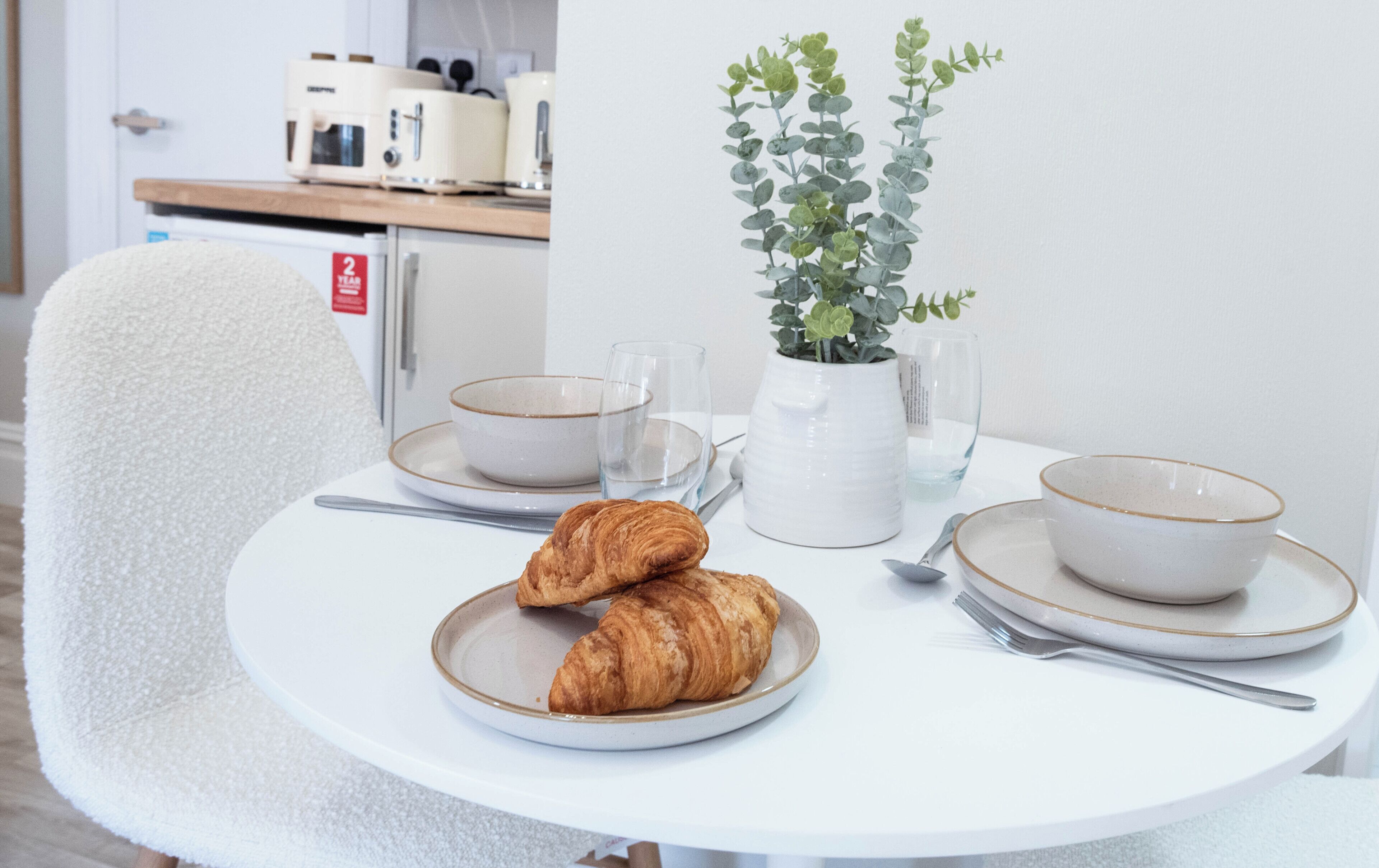 Ein runder Esstisch mit einem weißen Stuhl, zwei Schüsseln, zwei Weingläsern, einer Topfpflanze und zwei Croissants auf einem Teller.