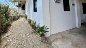 Un pasillo exterior cubierto con un sendero de grava, plantas en macetas y un edificio de fachada blanca y techo de madera.