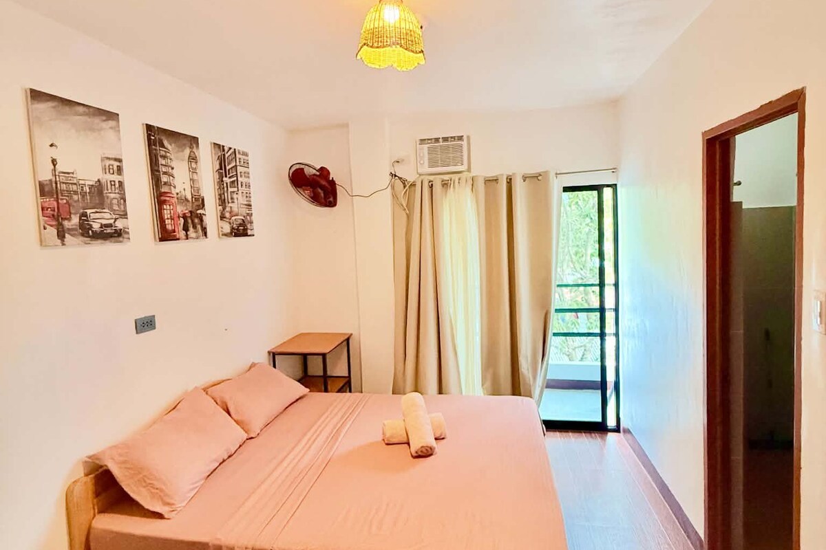 A bedroom with a bed, a bedside table, a lamp, a window with curtains, and wall art.