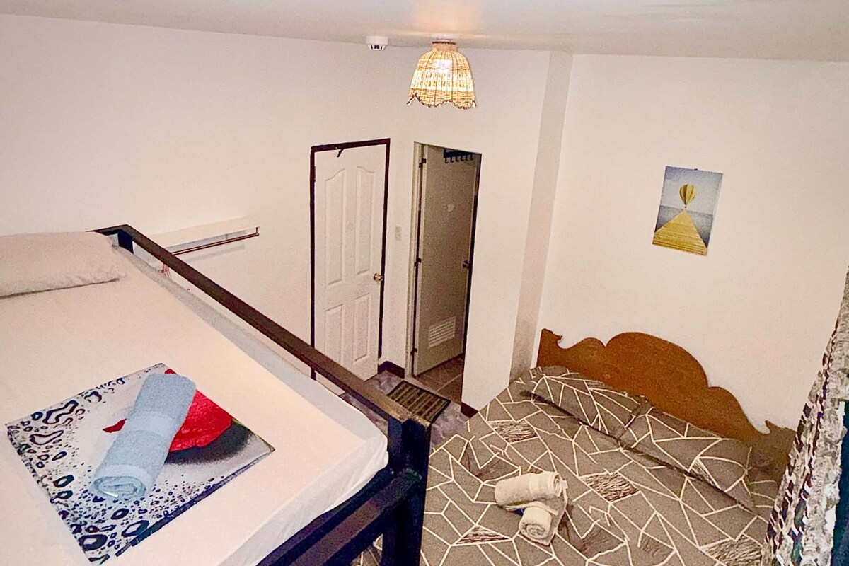 A room with a bed, a desk, and a chair. There is a door leading to another room.