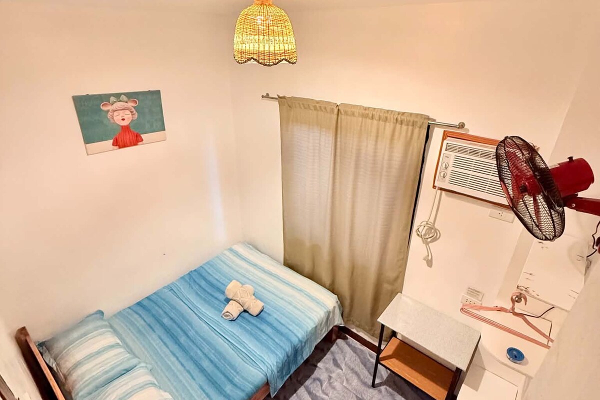 A small, single-bed room with a wooden frame, a blue-striped bedspread, a white wall with a picture, a ceiling light, a window with a curtain, and a wall-mounted fan.
