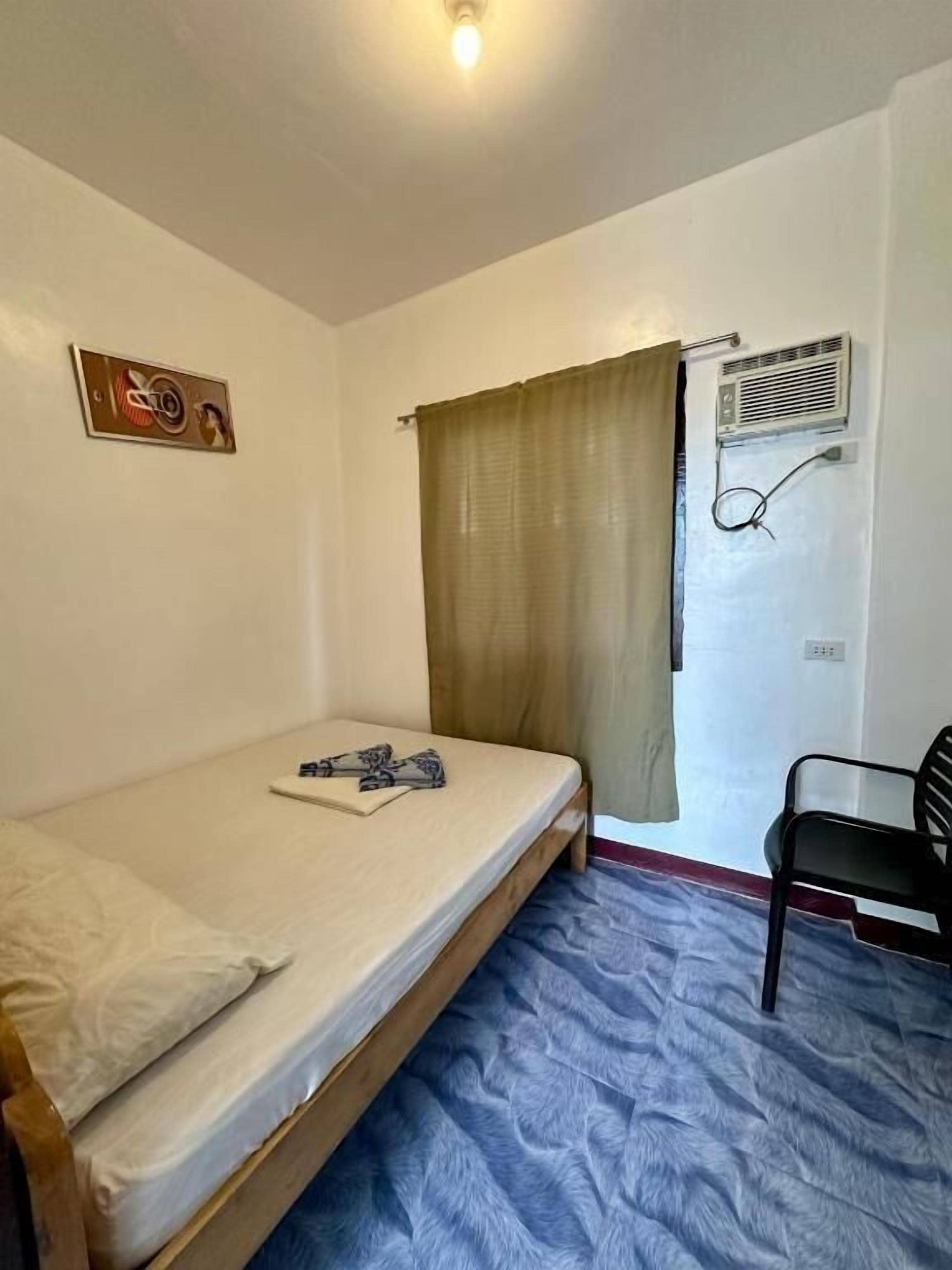 Ein kleines Zimmer mit einem Einzelbett, einem Stuhl, einem Fenster mit Vorhängen und einer Wand-Klimaanlage.