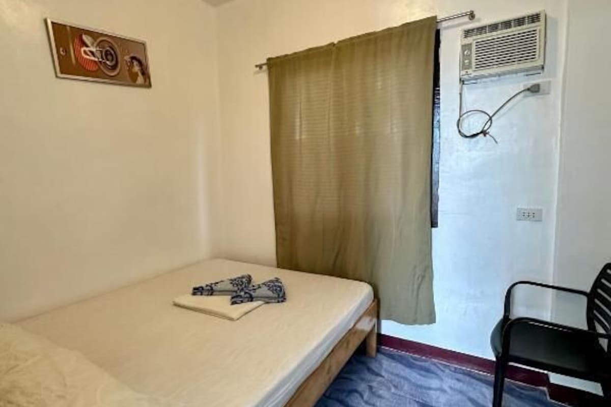 A small room with a single bed, a chair, a window with curtains, and a wall-mounted air conditioning unit.