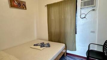 Ein kleines Zimmer mit einem Einzelbett, einem Stuhl, einem Fenster mit Vorhängen und einer Wand-Klimaanlage.
