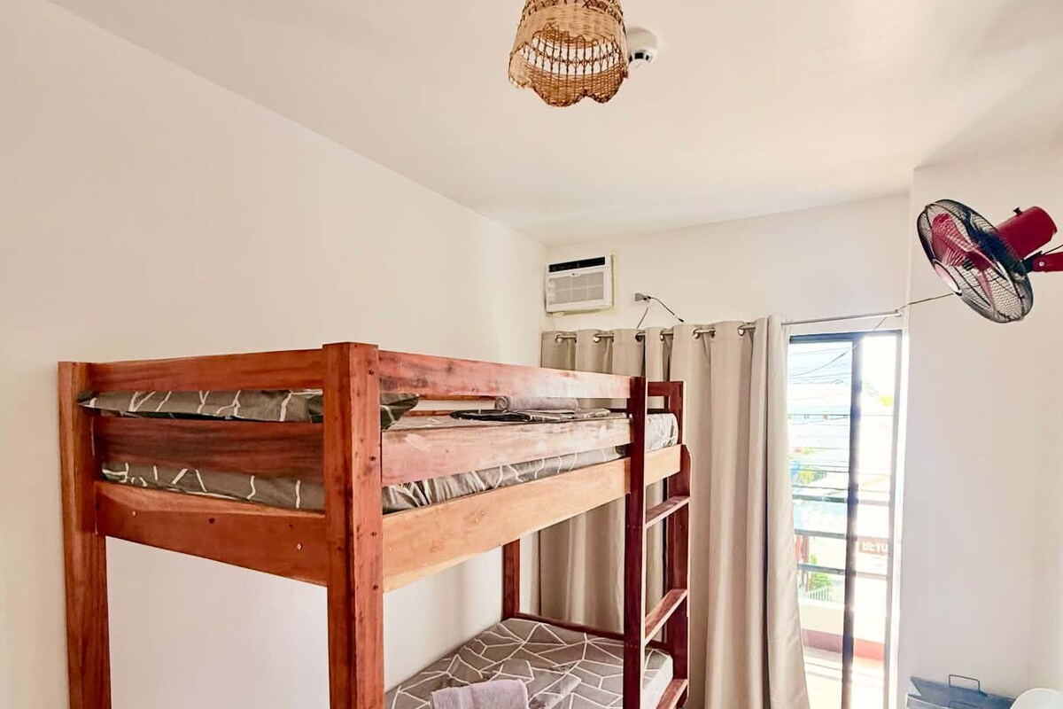 A bunk bed with a woven ceiling light fixture, a window with curtains, and a white plastic chair.