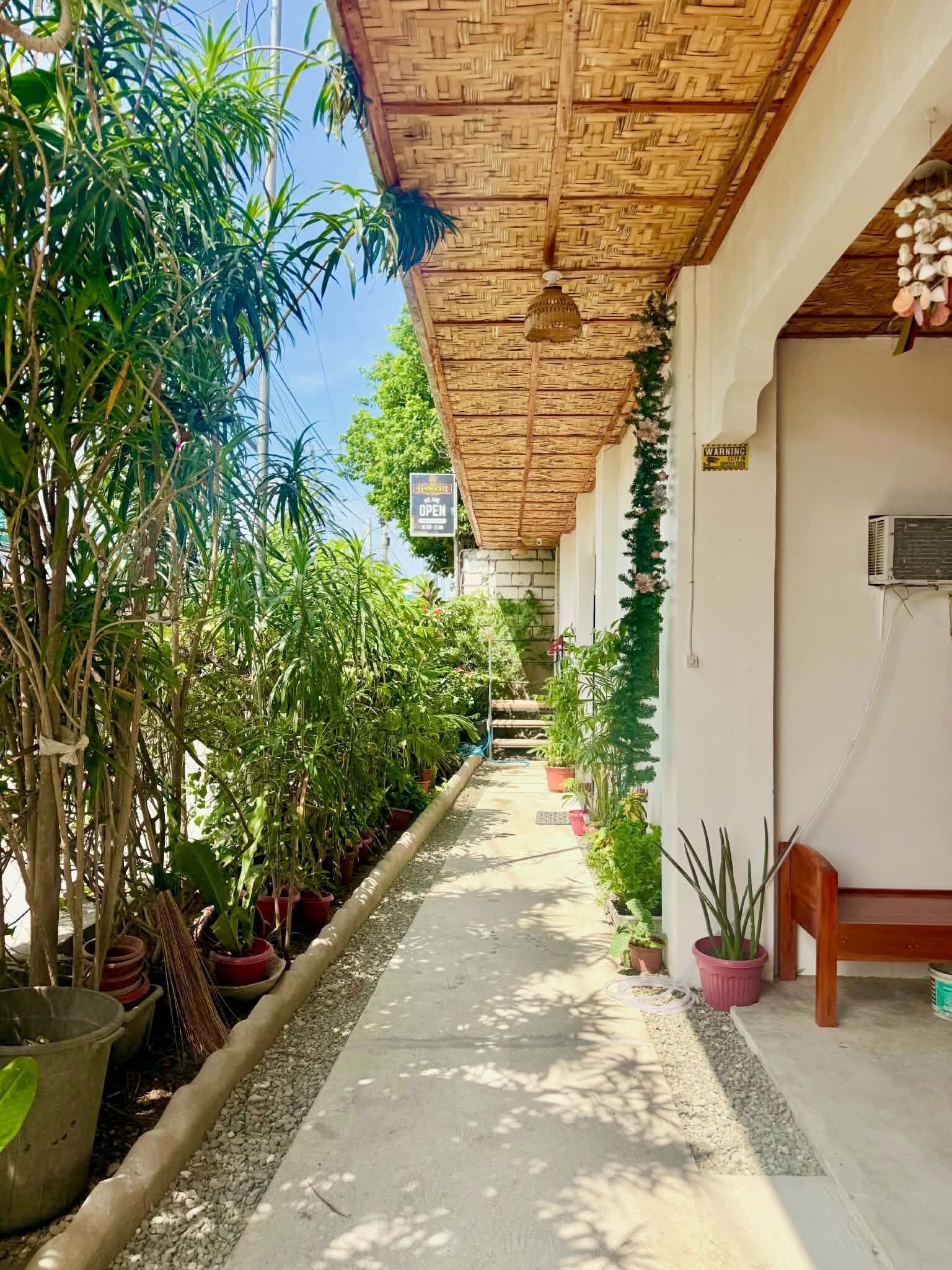 A covered walkway lined with potted plants, leading to a building with a woven ceiling.