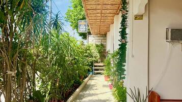 A covered walkway lined with potted plants, leading to a building with a woven ceiling.