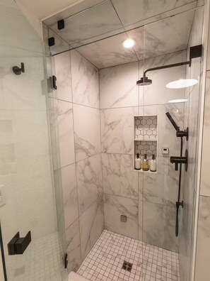 A modern shower room with a glass door, marble tiles, a handheld showerhead, and a built-in shelf with bottles. - The Studio - Downtown Jacksonville (Jacksonville)