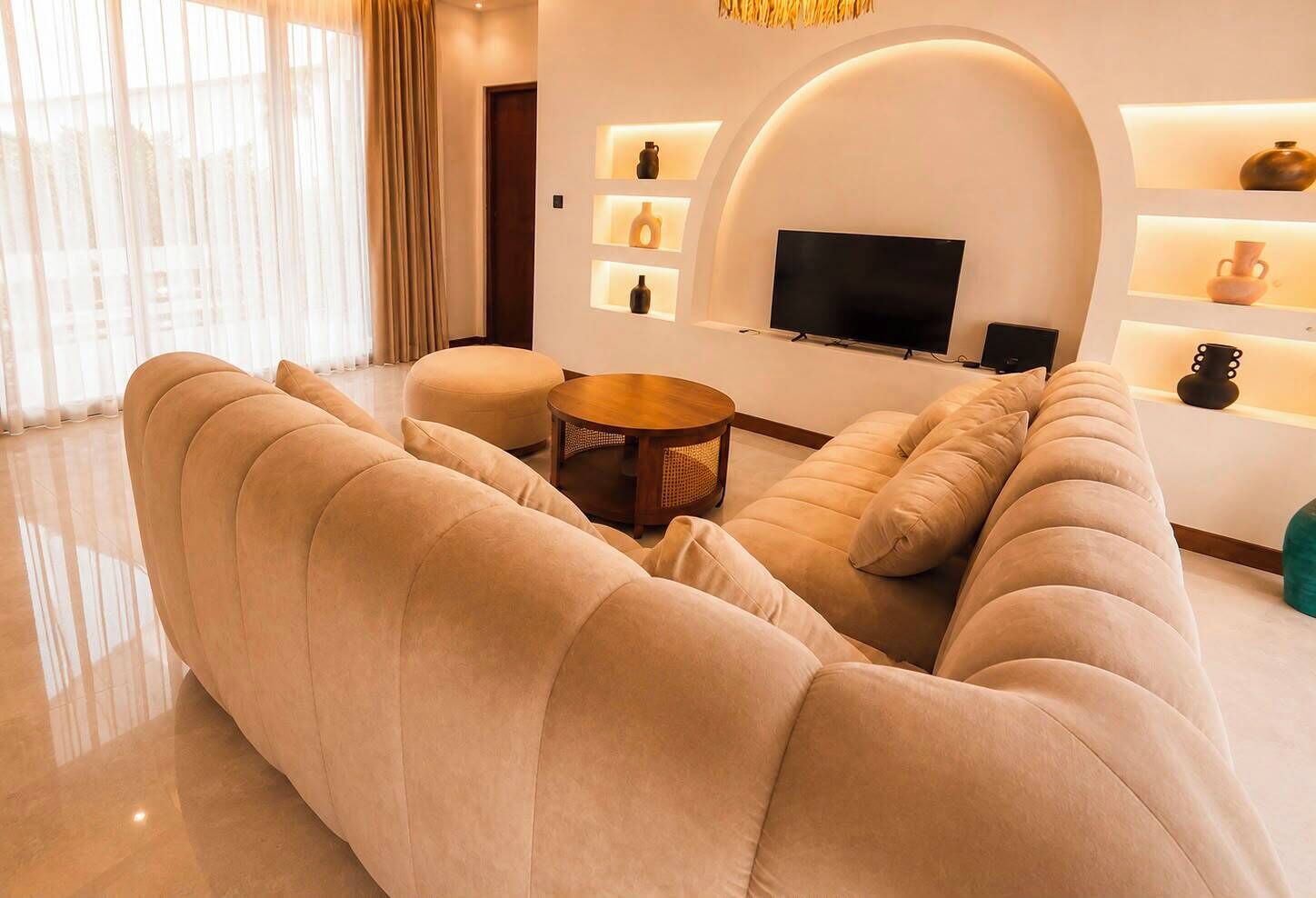 A modern living room with a large beige sofa, a round coffee table, and a flat-screen TV mounted on a white wall.