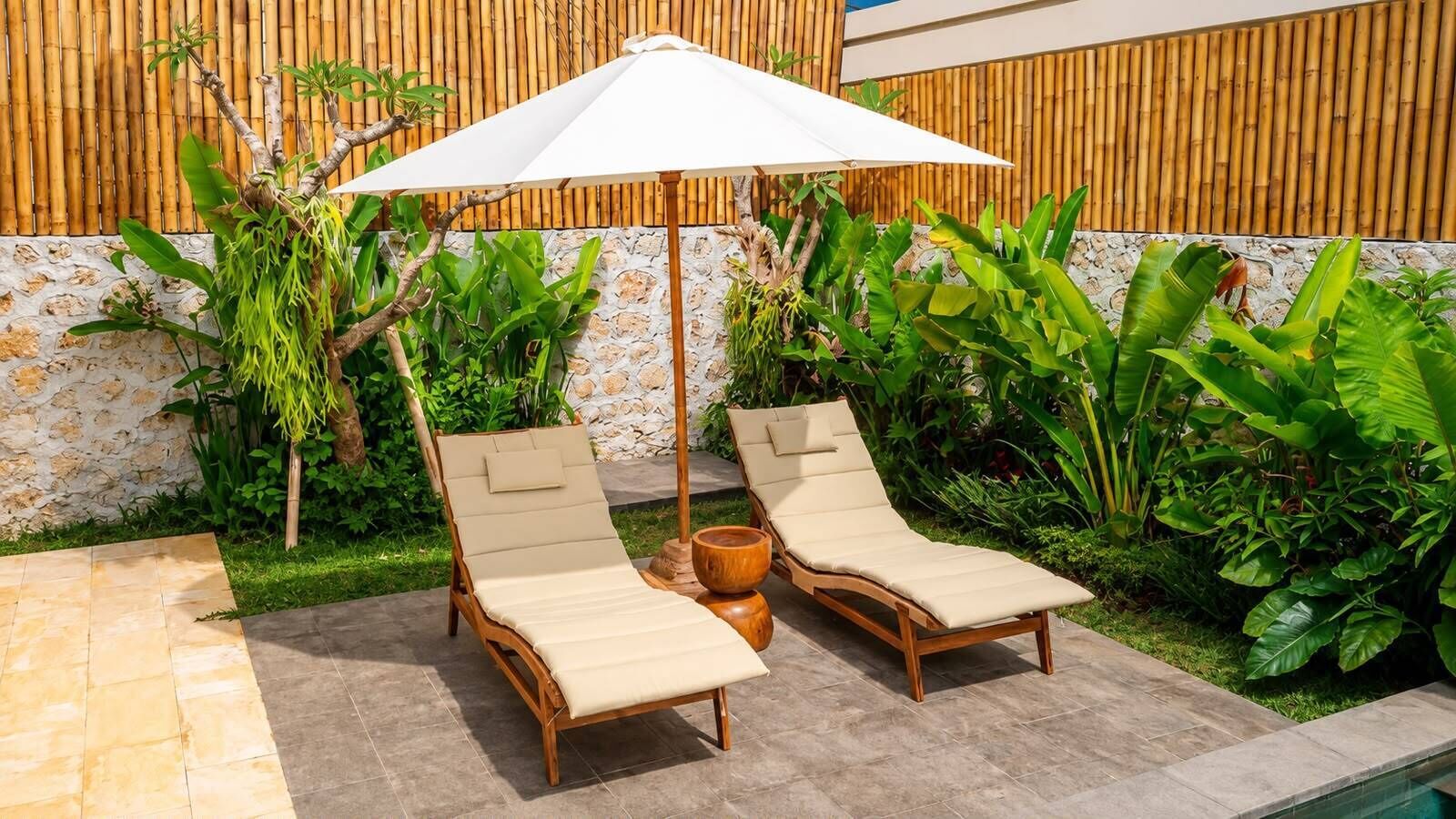 Two wooden lounge chairs with beige cushions and a white umbrella, positioned on a tiled patio with a bamboo wall and lush greenery in the background.