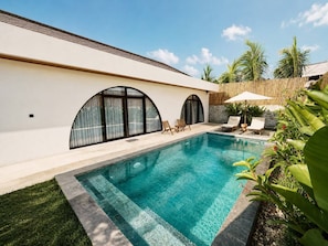 A swimming pool with a tiled edge, surrounded by a grassy area and a white building with arched windows. - Casa Anagata Pandawa (Badung)