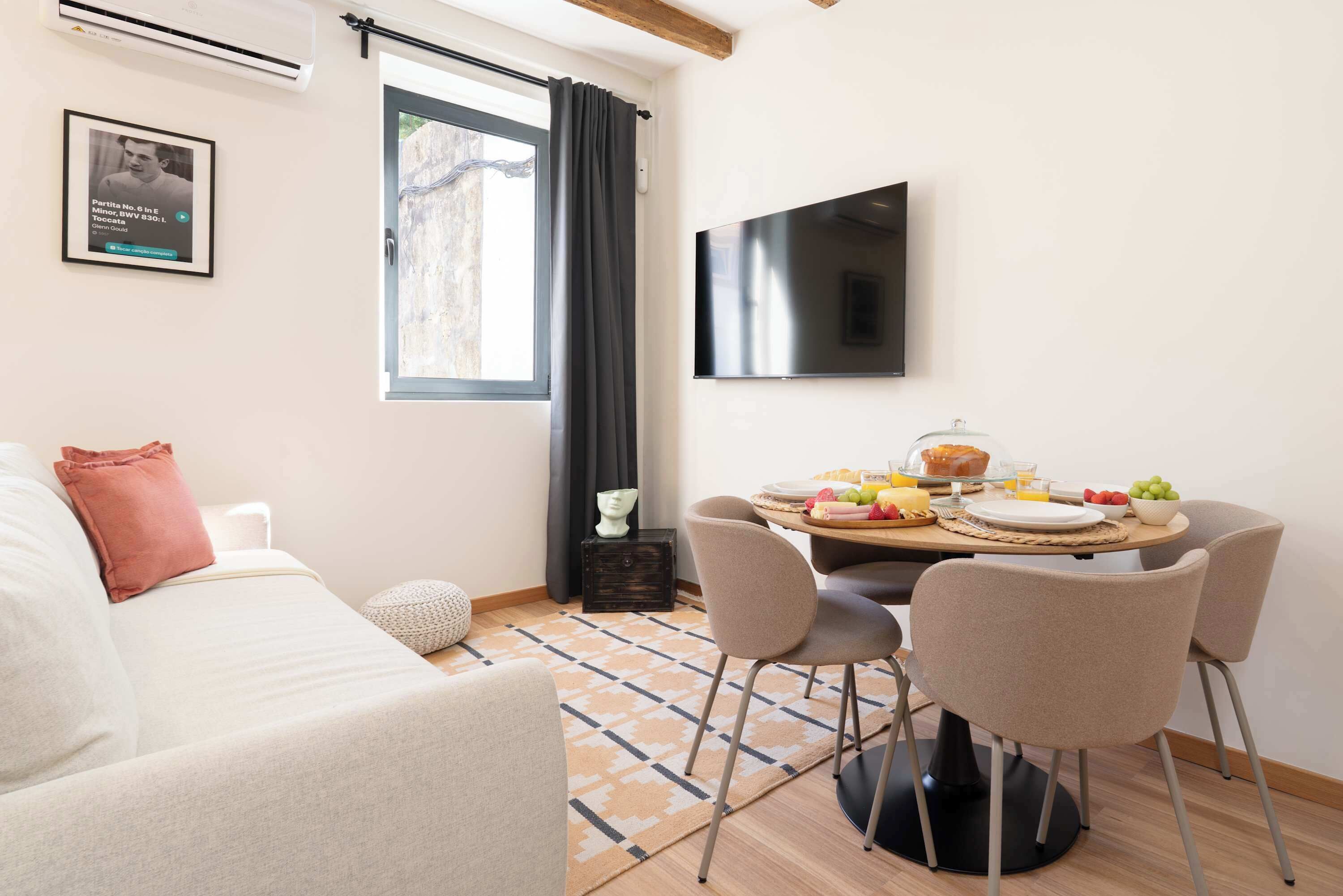 Una sala moderna con un sofá, una mesa de comedor lista para el desayuno, un televisor en la pared y un ventanal con cortinas.