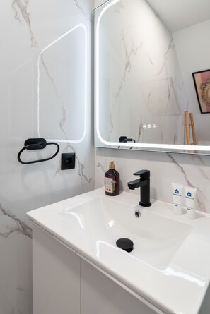 Un baño moderno con lavamanos de mármol blanco, grifo negro y un espejo grande con iluminación integrada.