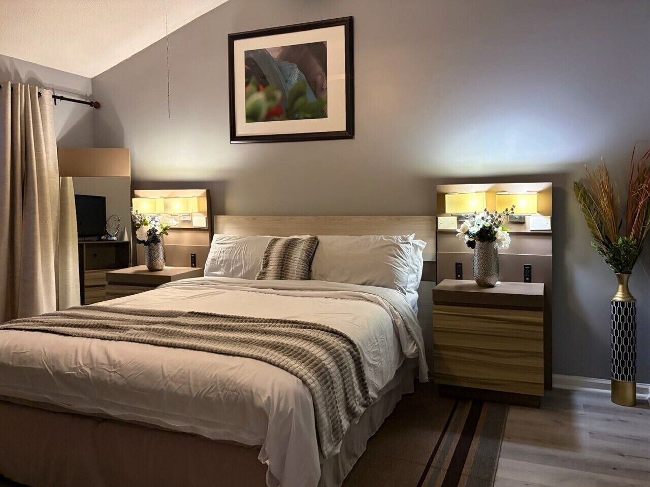 Un dormitorio moderno con una cama grande, mesitas de noche, una lámpara de noche, un cuadro enmarcado en la pared y un jarrón con flores.