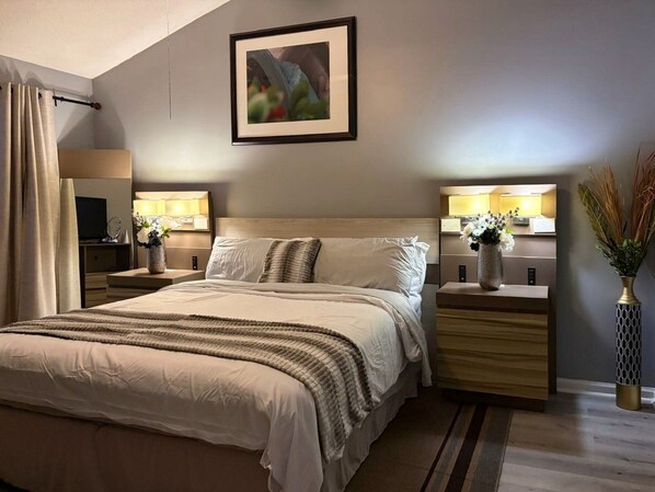 A modern bedroom with a large bed, bedside tables, a nightstand, a framed picture on the wall, and a vase with flowers.