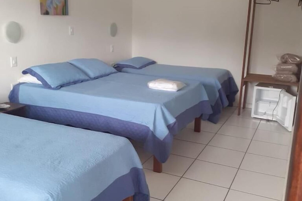 A hotel room with two beds, a small table, a mirror, and a shelf with bags.