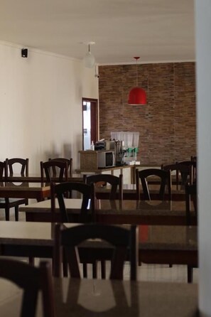 A dining area with wooden tables and chairs, a kitchen in the background, and a red pendant light. - HOTEL SOL A SOL (Cananéia)