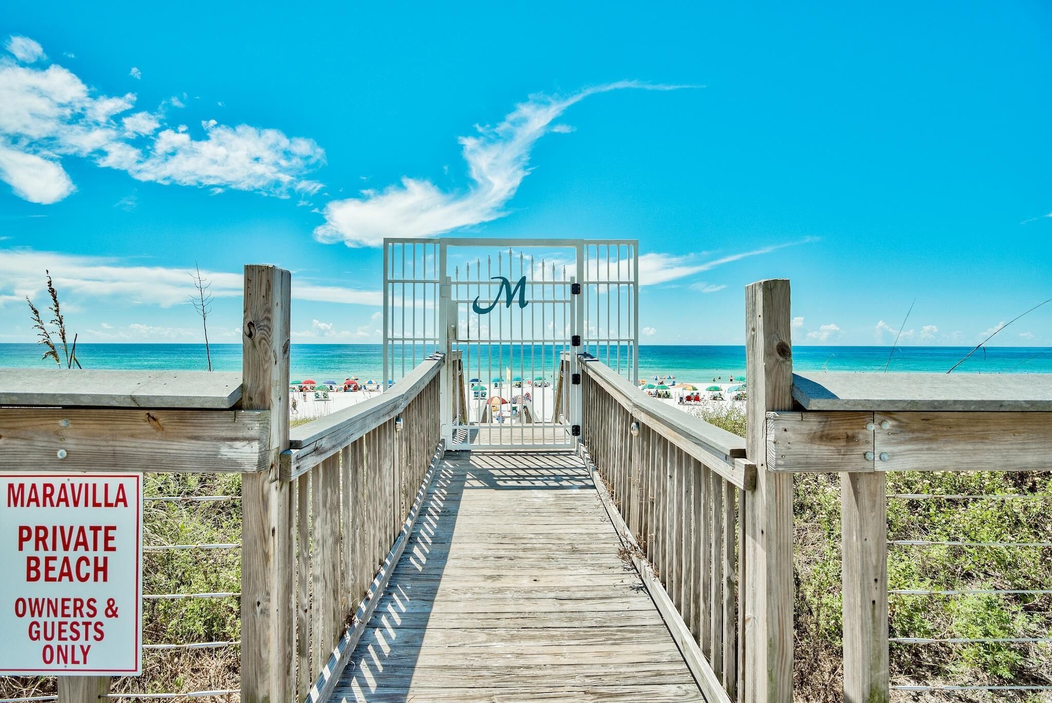 A wooden boardwalk leading to a private beach with a gate marked with a 'M'.