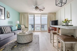A modern living room with a grey sofa, a round ottoman, a television, and a balcony with a view. - Pelican Beach Resort 904- Seaside Serenity (Destin)
