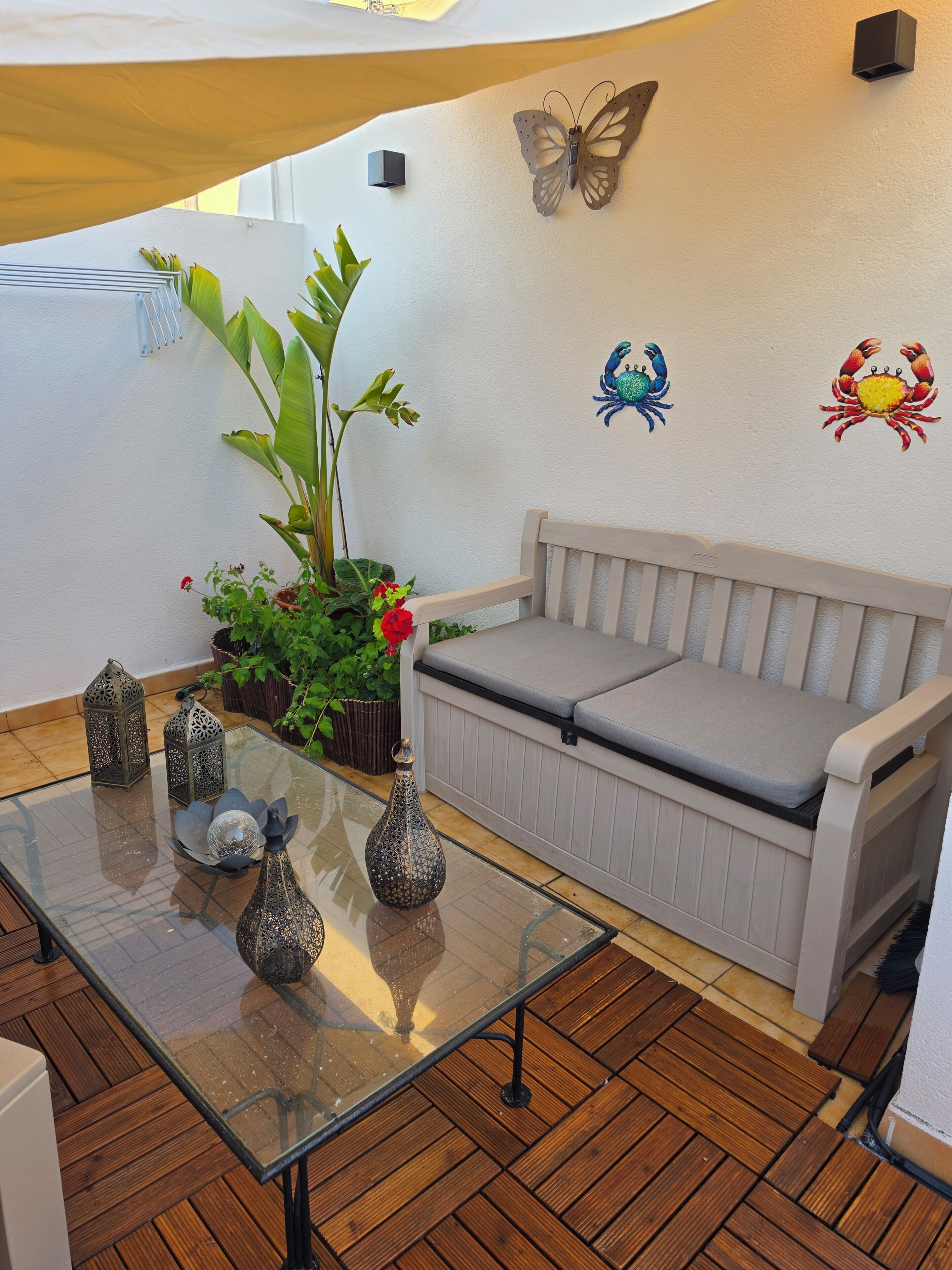 Una mesa con cubierta de vidrio, adornada con jarrones decorativos, un banco acolchado, plantas en macetas y detalles en la pared como una mariposa y cangrejos.