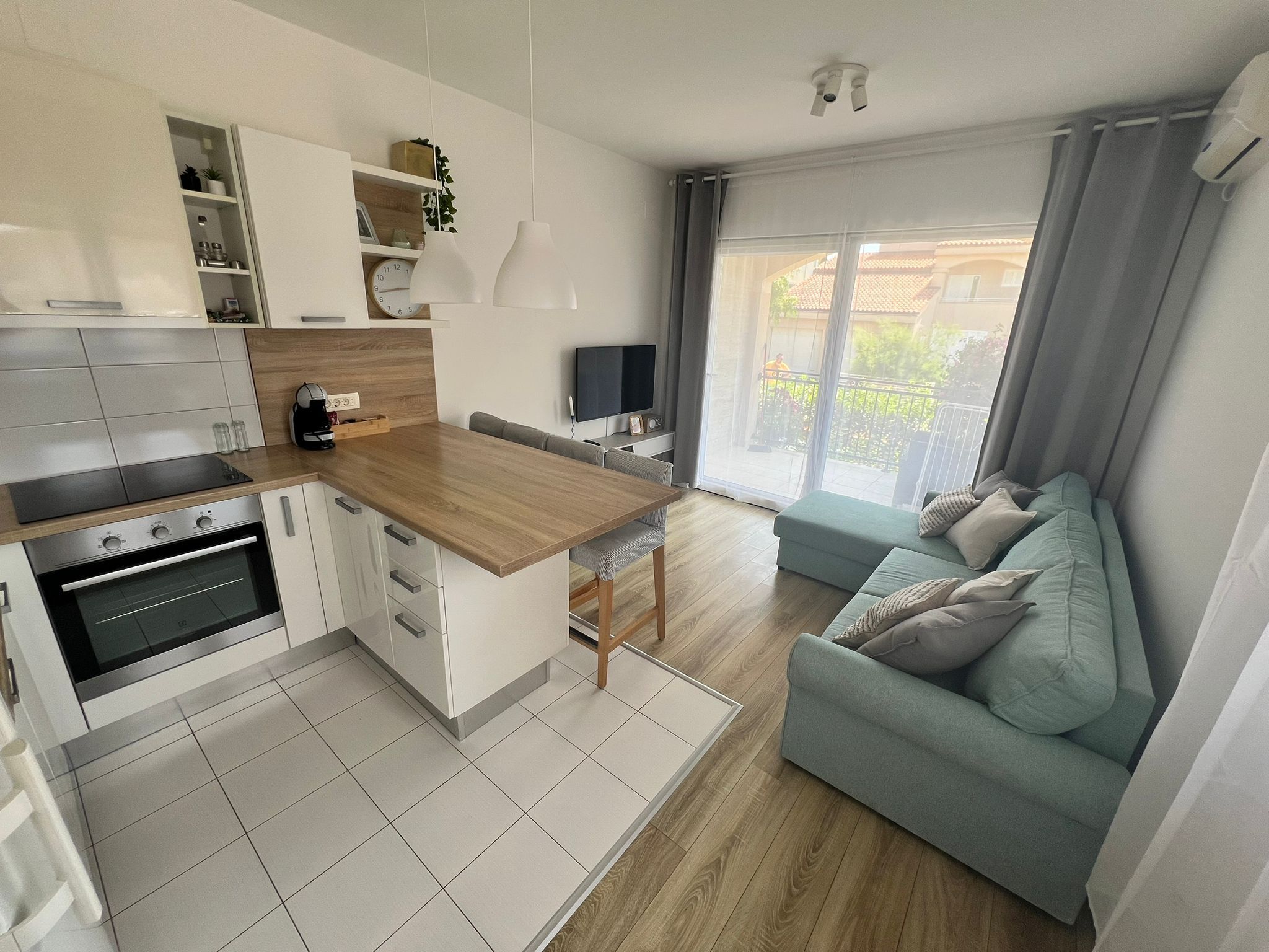 A modern kitchen with a built-in oven, a dining area with a wooden table and chairs, and a living room with a sofa and a TV.