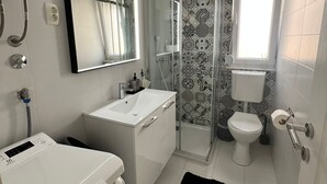 Casa de banho moderna com lavatório branco, espelho e sanita. Zona de duche com porta de vidro e um tapete preto no chão.