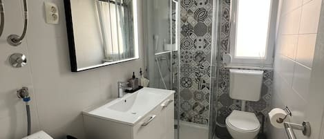 Casa de banho moderna com lavatório branco, espelho e sanita. Zona de duche com porta de vidro e um tapete preto no chão.