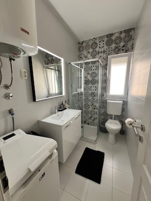 Een moderne badkamer met een witte wasbak, spiegel en toilet. Er is een douchegedeelte met een glazen deur en een zwart vloerkleed.
