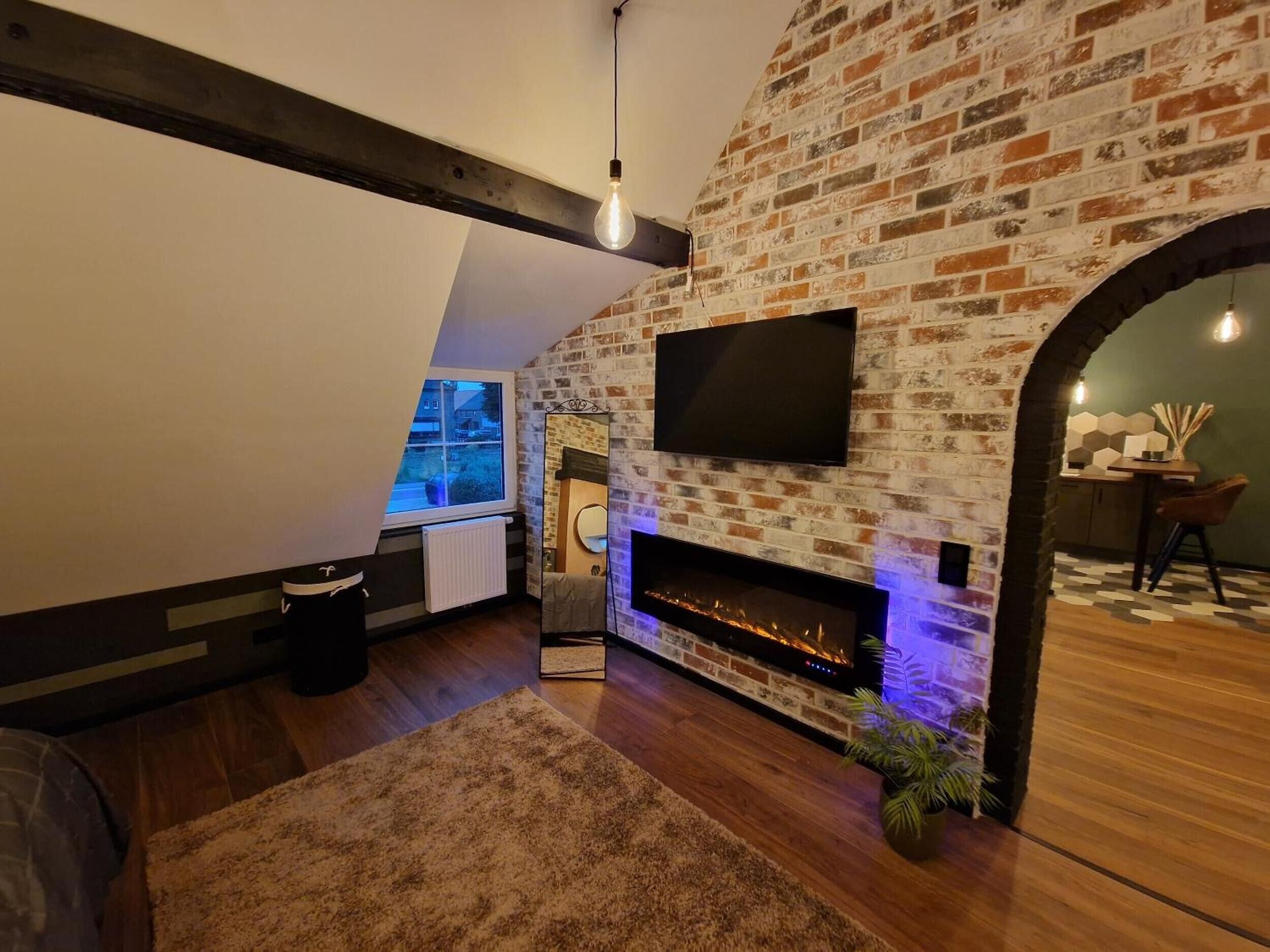 A modern living room with exposed brick walls, a flat-screen TV mounted above a fireplace, and a cozy seating area with a rug and a plant.