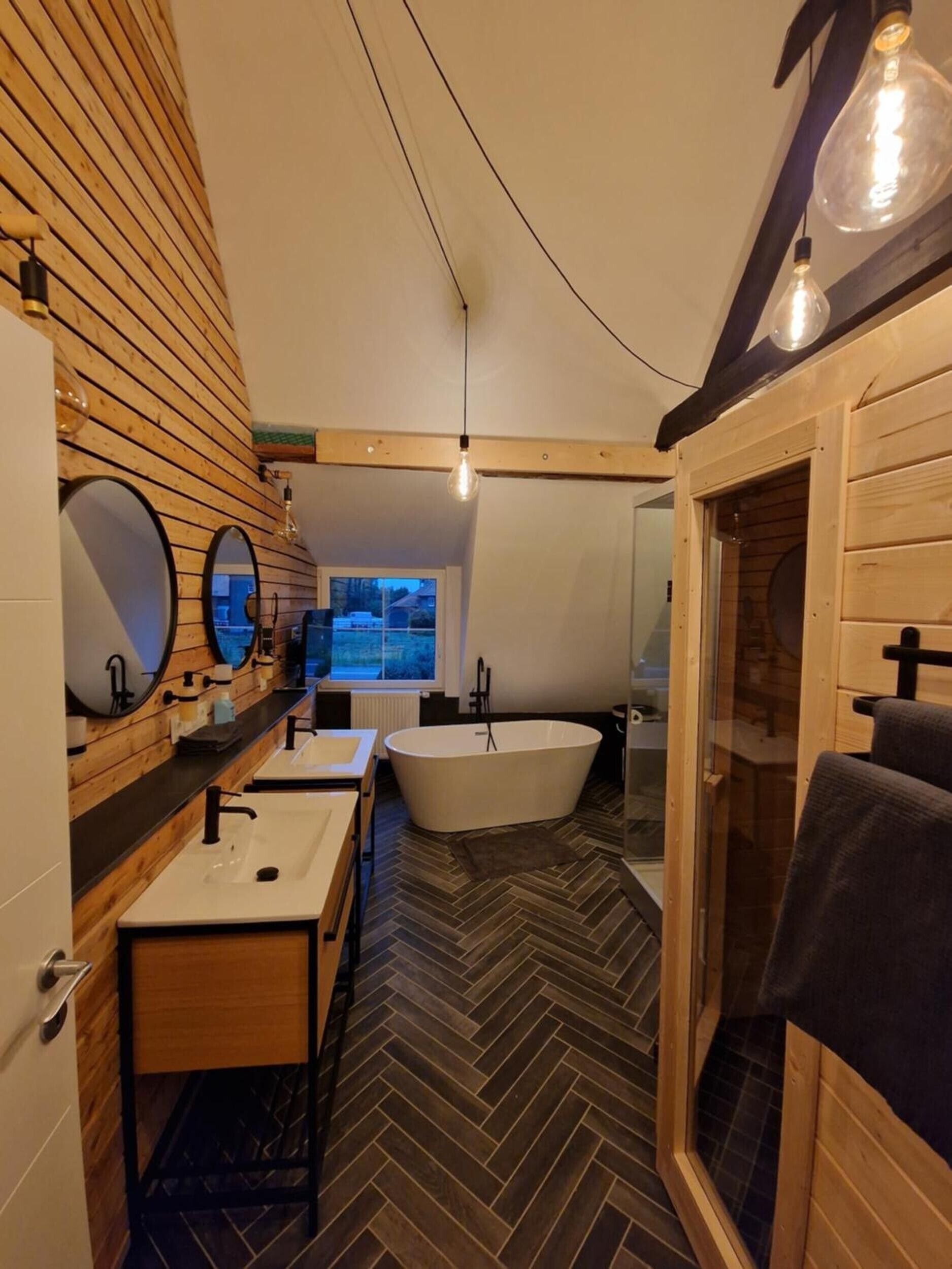 Een moderne badkamer met een visgraatvloer, houten lambrisering, een vrijstaand bad en een grote spiegel.