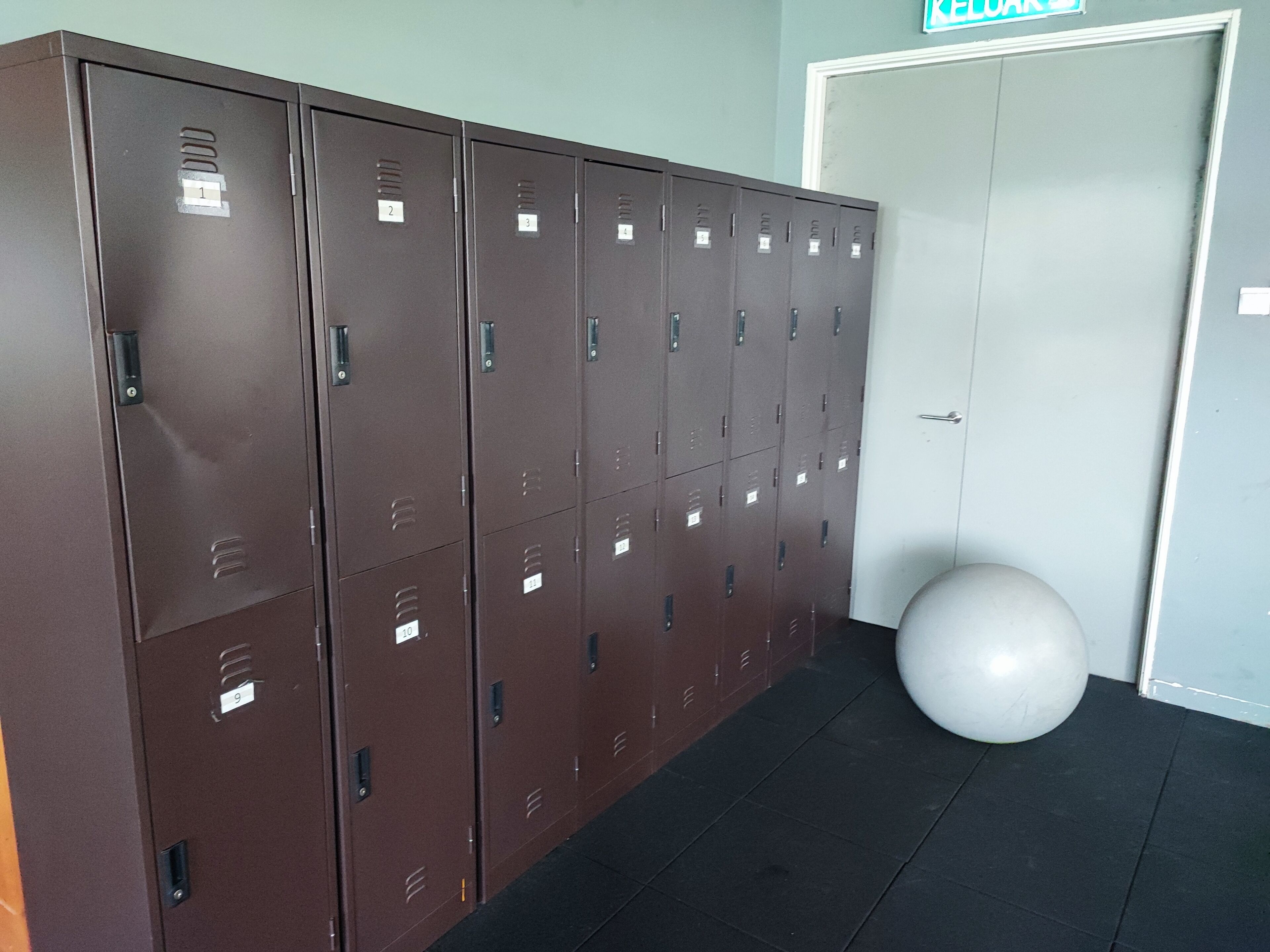 A row of brown lockers with numbered labels, a white exercise ball, and a door with a sign above it.