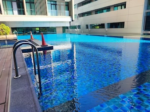 Een modern zwembad met helderblauw water, een houten terras en een modern gebouw op de achtergrond.