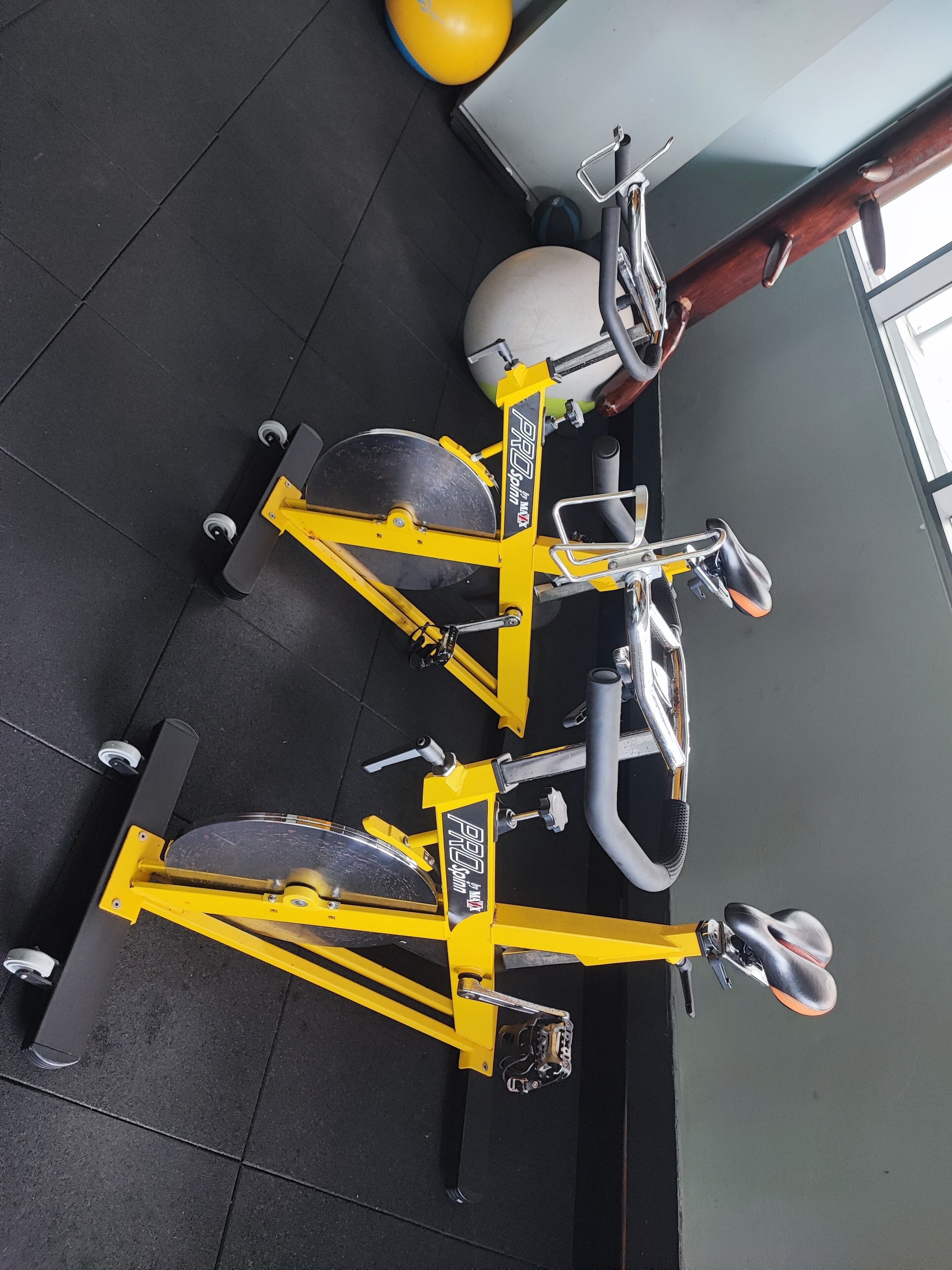Un vélo d’appartement et un appareil de rame dans une salle de sport, avec un sol noir et une fenêtre en arrière-plan.