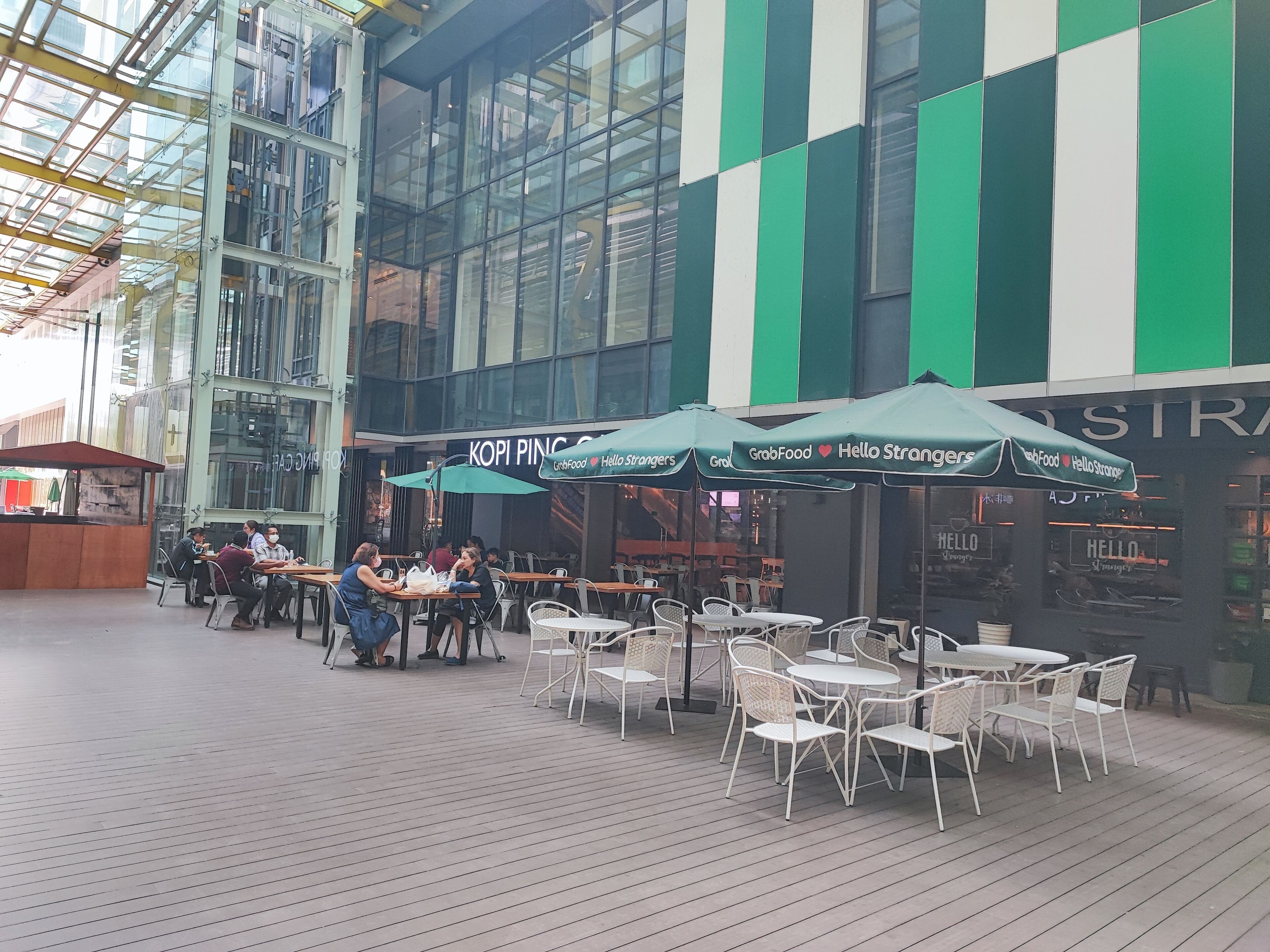 An outdoor dining area with tables and chairs, a glass building in the background, and umbrellas with text.