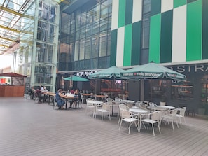 Een buitenterras met tafels en stoelen, een glazen gebouw op de achtergrond en parasols met tekst.