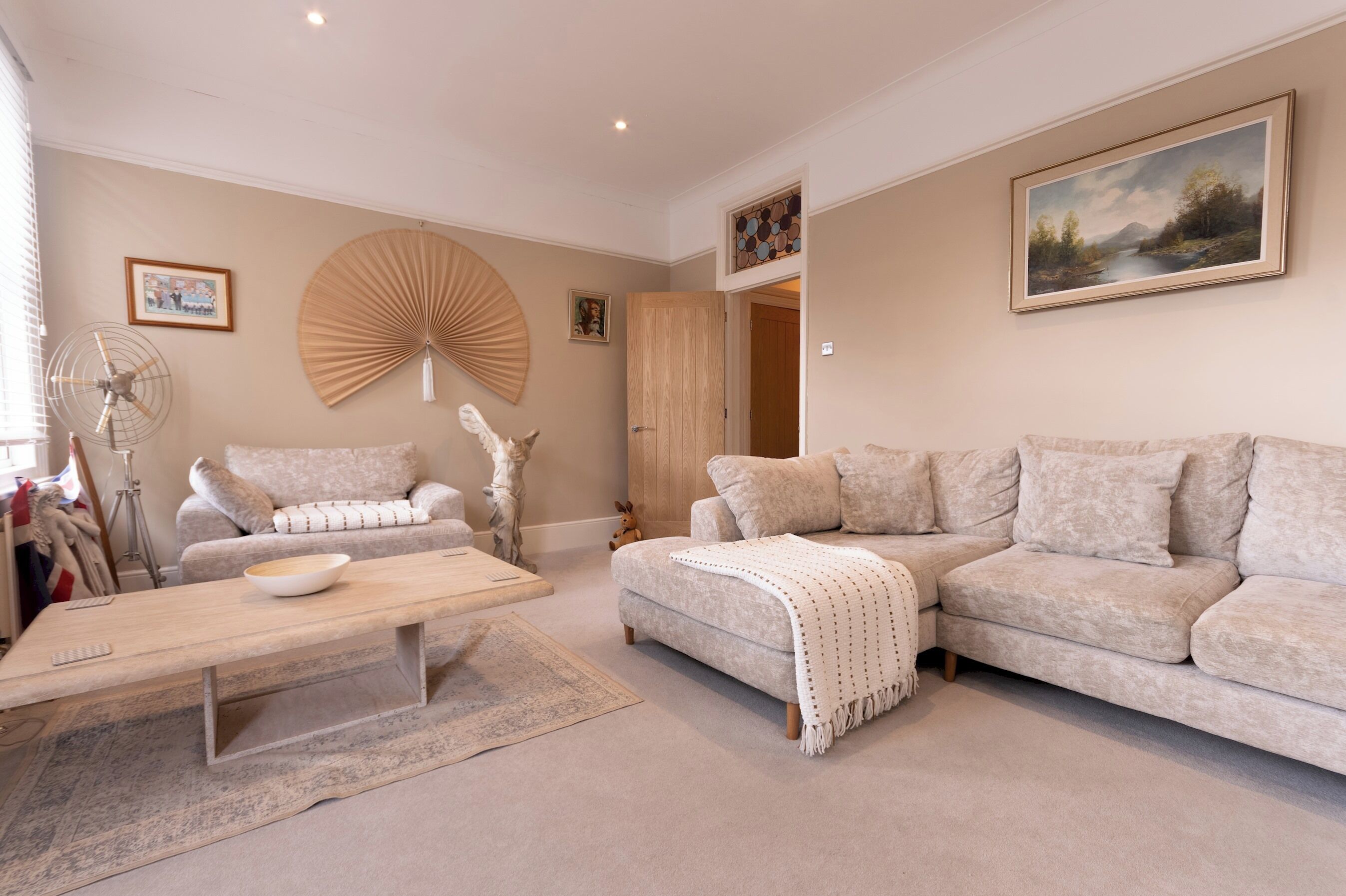 A living room with a beige sofa, a coffee table, a fan, and a painting on the wall.