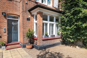Une maison en briques avec une porte d’entrée sombre, des volets blancs et un petit jardin agrémenté de plantes en pot.
