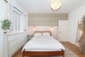 Ein Schlafzimmer mit einem Bett, einem Nachttisch, einer Heizung, einem Fenster mit Rollläden und einer Wand mit einem Blattmuster.