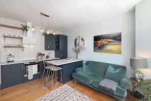 Eine moderne Küche mit Fischgrätmuster-Fliesen an der Rückwand, einer zentralen Kücheninsel, Barhocker und einem dunkelblauen Sofa in einem Wohnbereich mit einem Gemälde an der Wand.