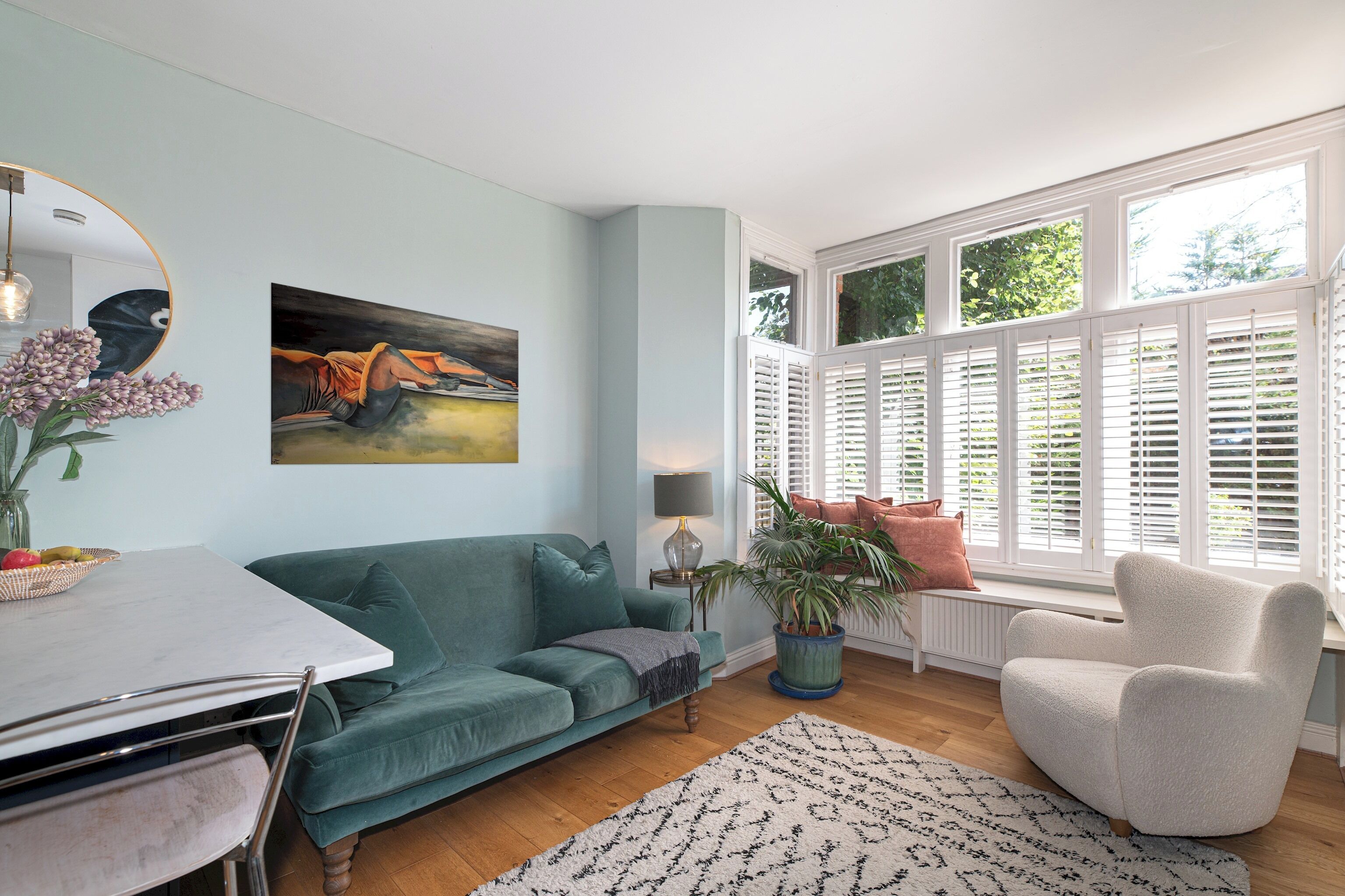 Un salon avec un canapé de couleur turquoise, un fauteuil blanc, une table à manger et une grande fenêtre avec des volets.