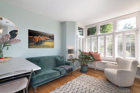 Una sala de estar con un sofá azul turquesa, un sillón blanco, una mesa de comedor y un ventanal con contraventanas.
