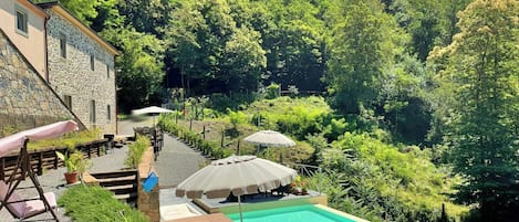 Un área de piscina con un edificio de piedra, mobiliario exterior y abundante vegetación.